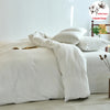 A white 100% cotton yarn-dyed duvet cover displayed on a bed with pillows and a wooden shelf in the background.