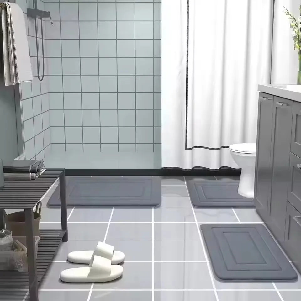 A collection of gray memory foam bathroom mats and white foam slippers are displayed on a tiled floor.