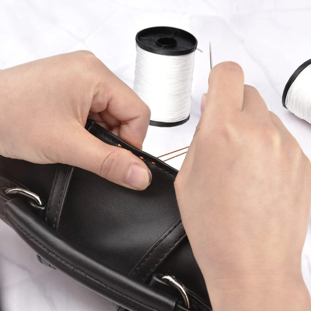 Hands are shown using a sewing needle and thread to work on a black leather item.