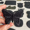 A hand holds a black embroidered leather butterfly patch with a self-adhesive backing.