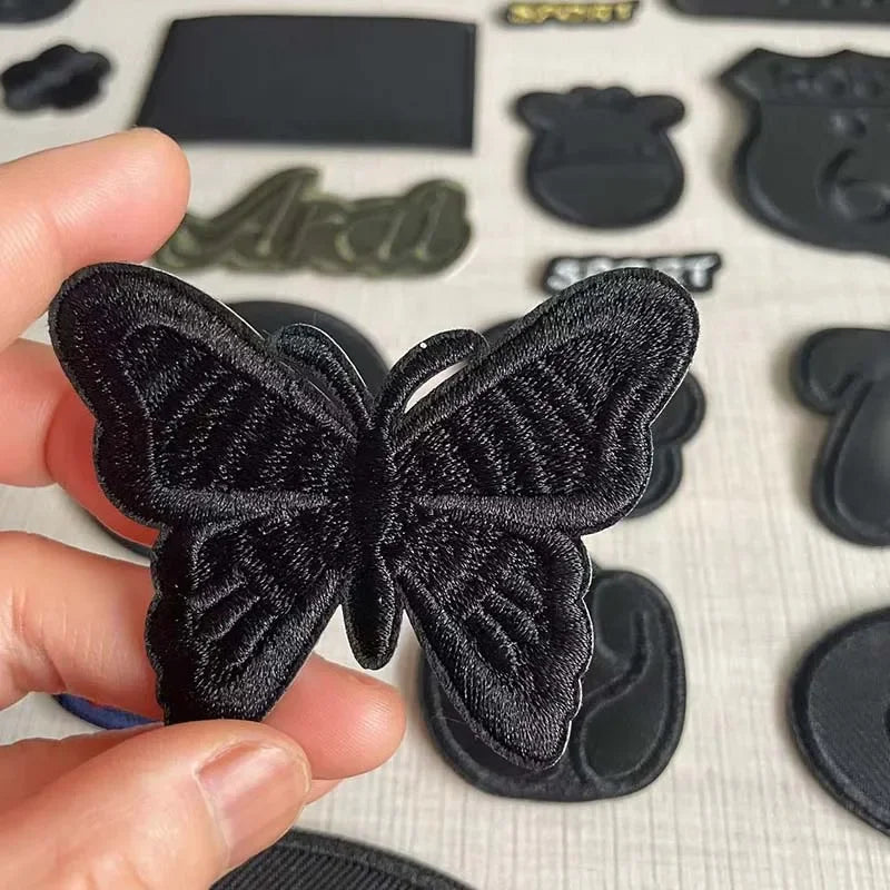 A hand holds a black embroidered leather butterfly patch with a self-adhesive backing.