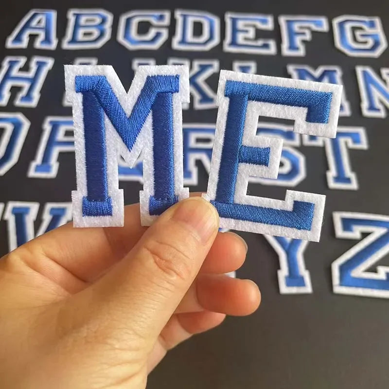 A hand holds an embroidered letter M and E from an alphabet set on a dark background.