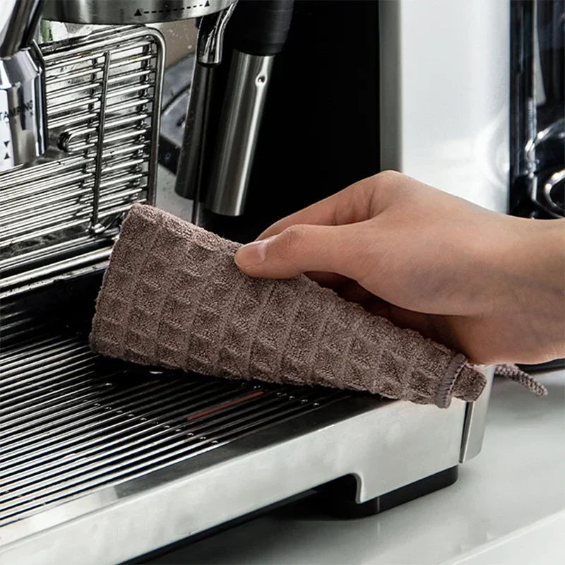 A hand is placing a brown microfiber cleaning towel onto the drip tray of a stainless steel coffee machine.