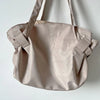 A light beige foldable tote bag with shoulder straps is shown against a white background.