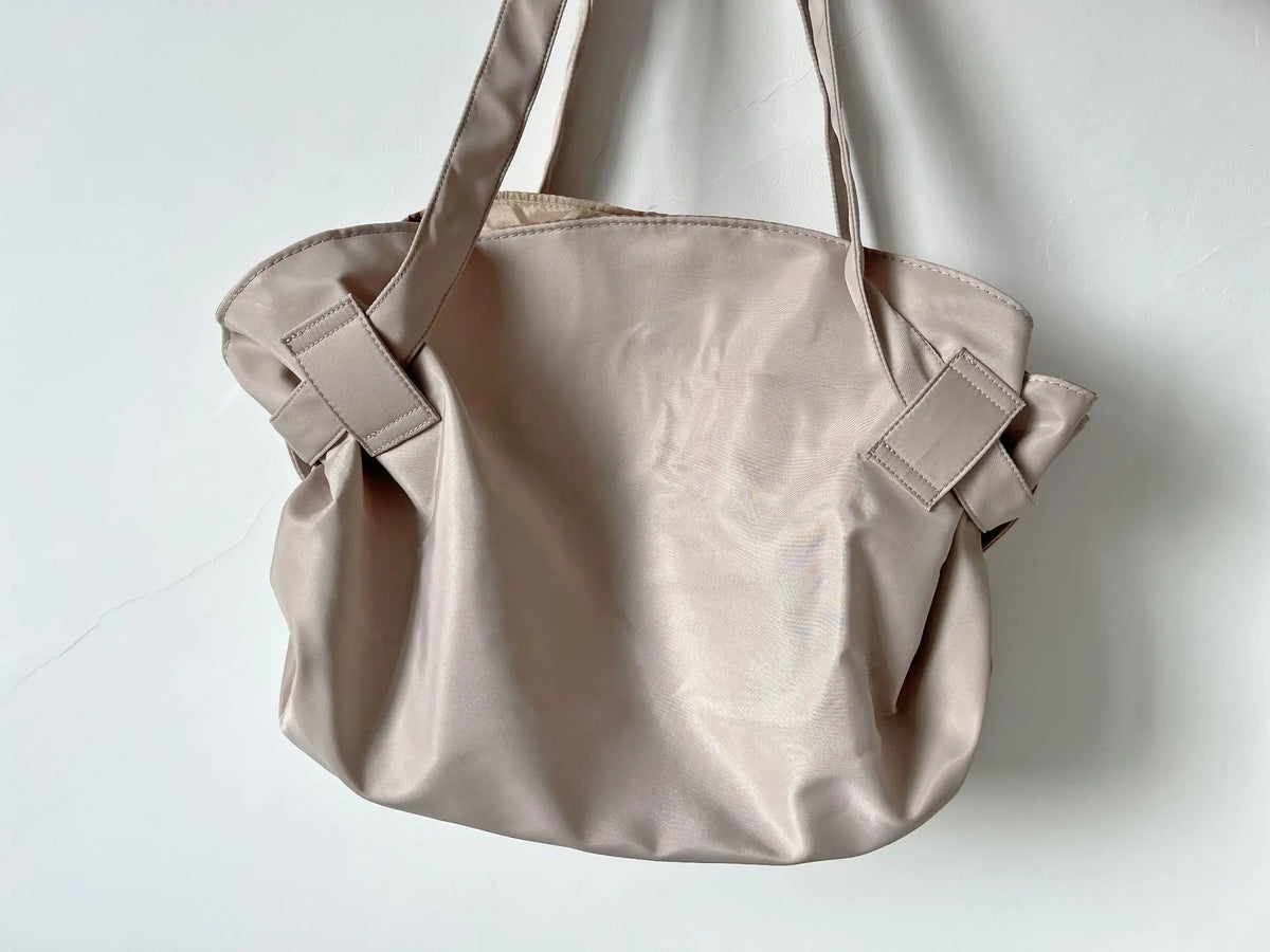 A light beige foldable tote bag with shoulder straps is shown against a white background.