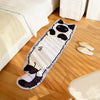 A long, rectangular white and black cartoon cat plush rug lying on a wooden floor next to a bed.