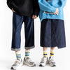 A person wearing dark blue jeans and a light blue sweater is standing, holding a pair of men's retro striped crew socks.
