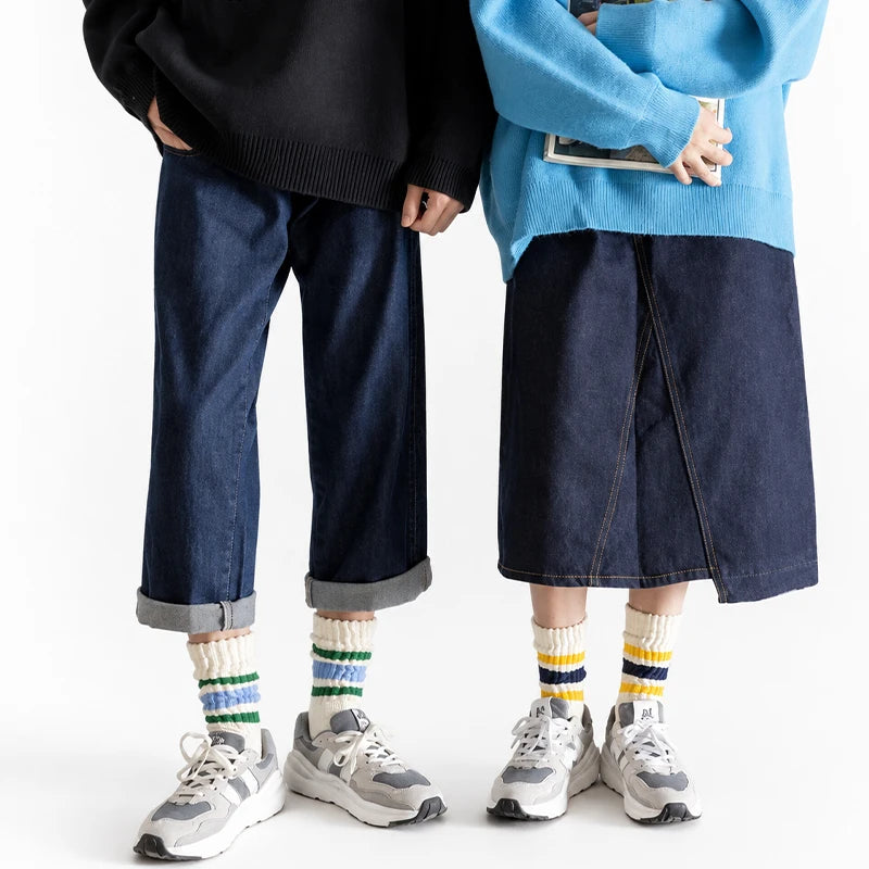 A person wearing dark blue jeans and a light blue sweater is standing, holding a pair of men's retro striped crew socks.