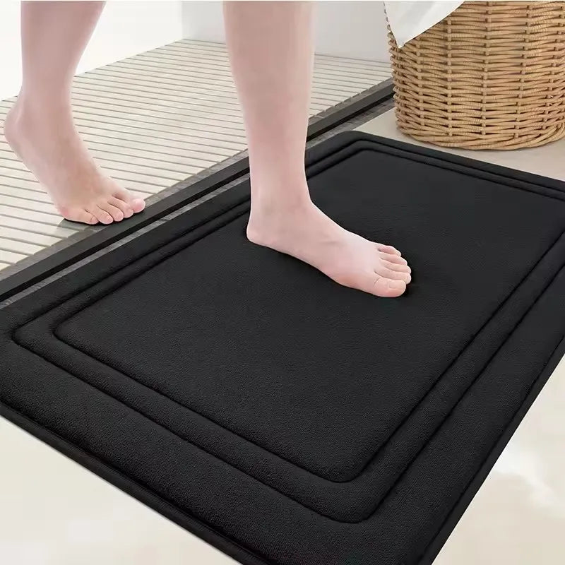 A black ultra soft memory foam bathroom mat is placed on a tiled floor with bare feet visible.
