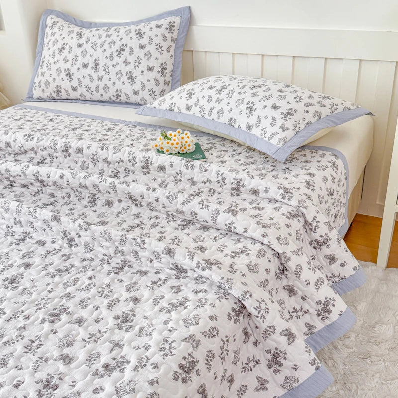 A white and gray floral quilted bedspread with light blue trim and matching pillows on a bed.