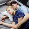 A woman is resting on a gray surface while a man gently rests his head on her shoulder.