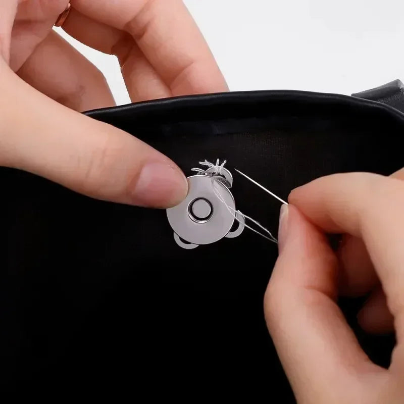 Hands are using a needle to attach magnetic snap fasteners onto a black bag or purse.