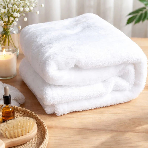 A stack of fluffy white flannel blankets resting on a wooden surface next to a glass candle and a brush.