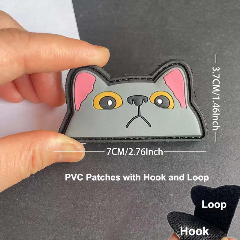 A hand holds a gray PVC patch shaped like a cat face with pink ears and yellow eyes, showing the hook and loop backing.