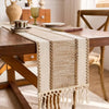 A beige macrame tassel table runner is draped across a rustic wooden dining table.
