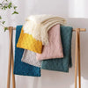 A display of oversized knitted tassel throw blankets in various shades including yellow, pink, teal, and cream.