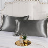 A set of dark gray satin pillowcases displayed on a white bed with a light upholstered headboard.