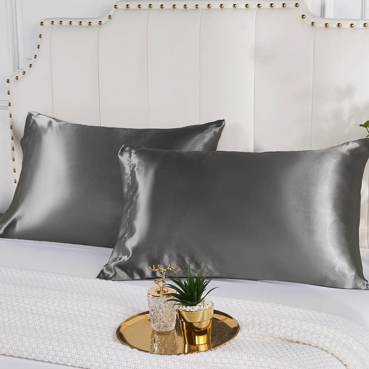 A set of dark gray satin pillowcases displayed on a white bed with a light upholstered headboard.