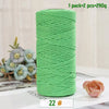 A spool of medium-thick, bright green hand-woven cotton yarn for crafting, next to a pink rose.