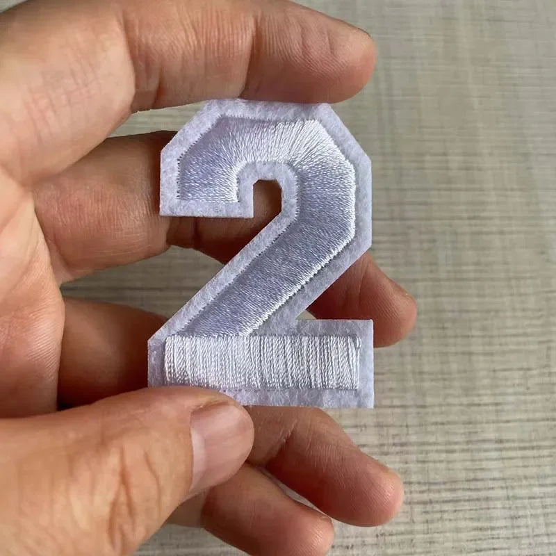 A hand holds a light purple embroidered number 2 patch for apparel.