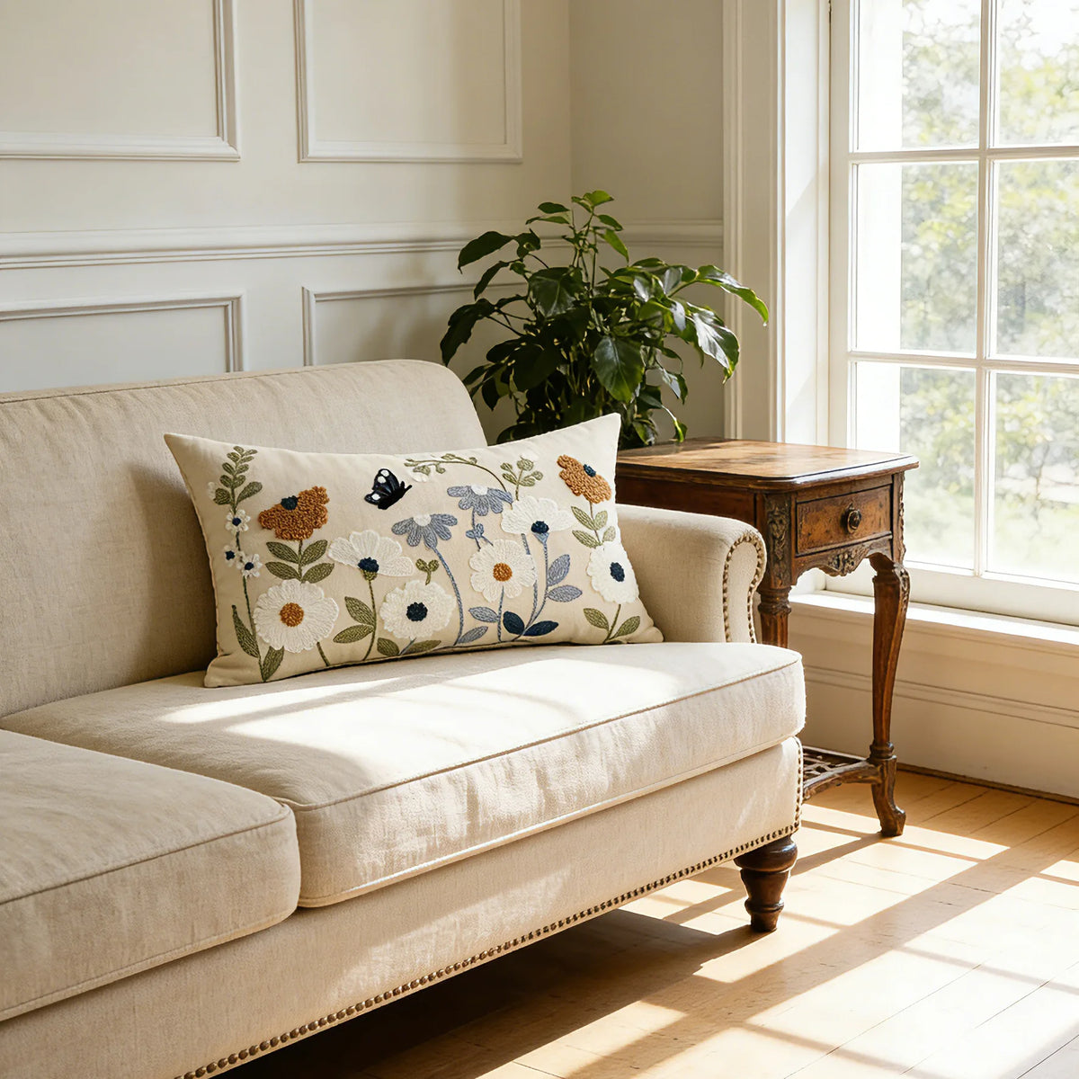 A beige sofa with a floral embroidered throw pillow on it, next to a wooden table and a large window.
