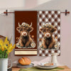 A set of two brown plaid kitchen towels featuring a cow motif, displayed on a wall.