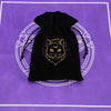 A black velvet pouch with a gold-colored cat face design rests on a purple surface.