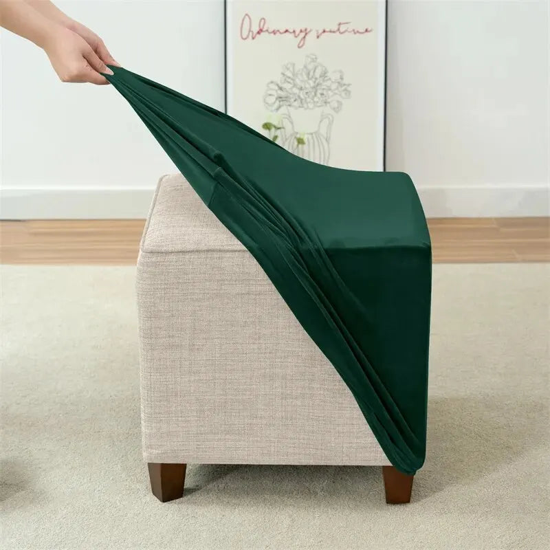A hand is draping a dark green velvet ottoman slipcover over a light beige upholstered furniture piece.
