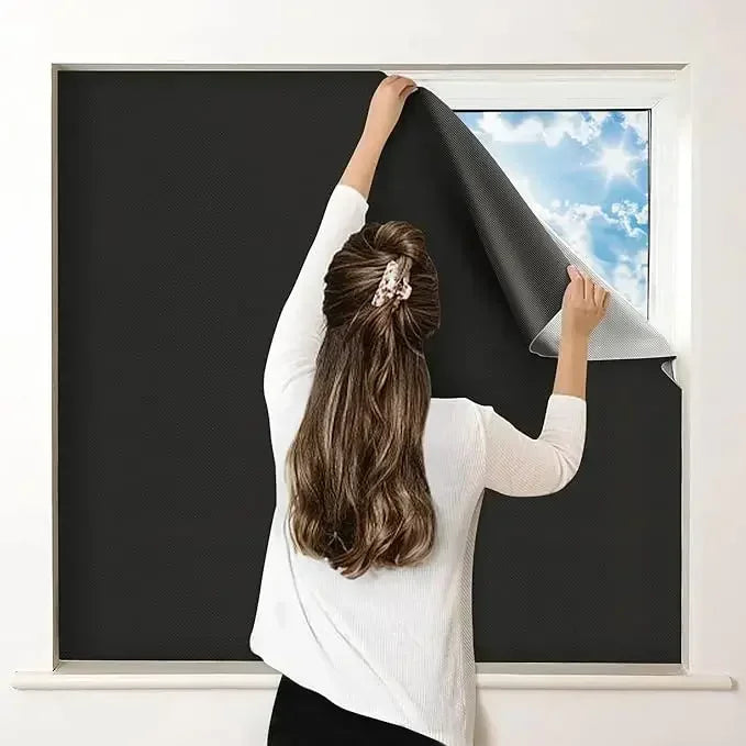A person is pulling down a black blackout window shade to reveal the bright blue sky outside.