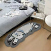 A gray and white cartoon cat plush rug is placed on a light wood floor next to a bed.