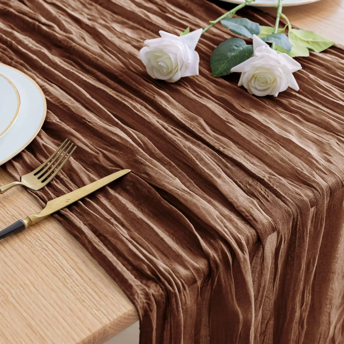 A brown cheesecloth table runner with draped folds, featuring white roses and gold cutlery on a wooden table.