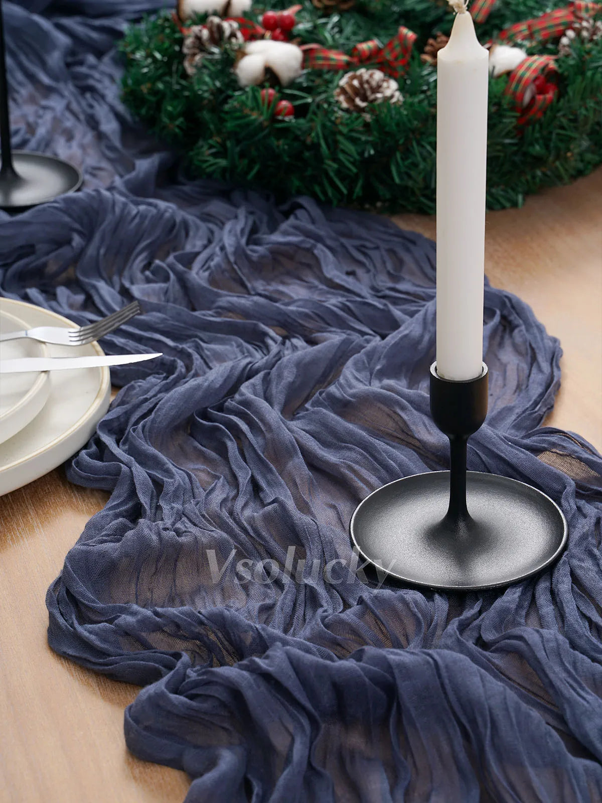 A dark blue crinkled cheesecloth table runner draped on a wooden surface with a lit white candle and greenery.