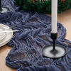 A dark blue crinkled cheesecloth table runner draped on a wooden surface with a lit white candle and greenery.