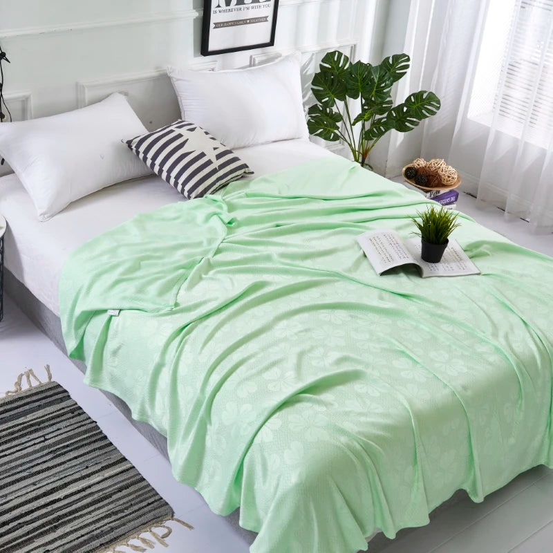 A light green bamboo fiber cooling blanket draped over a white bed with pillows and greenery.