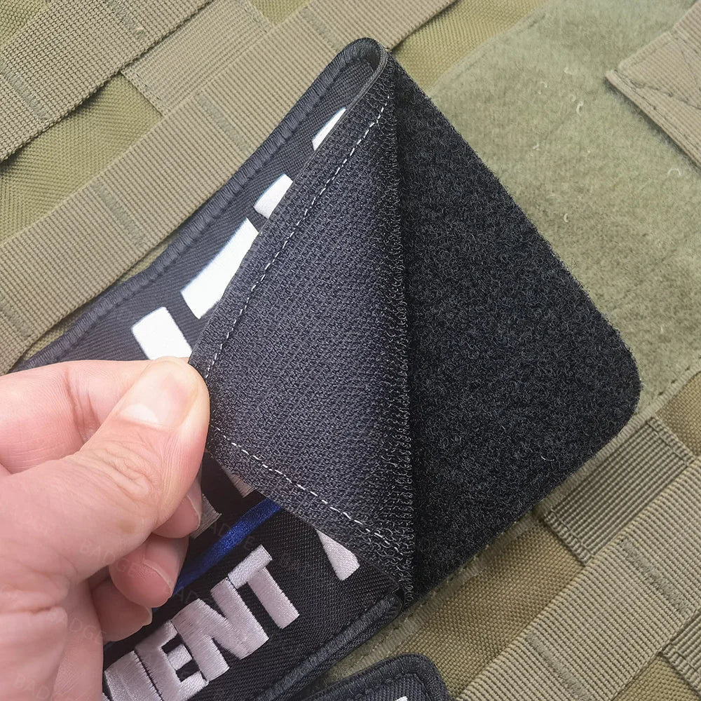 A close-up of a black tactical embroidered security patch being held by a hand against a green uniform background.