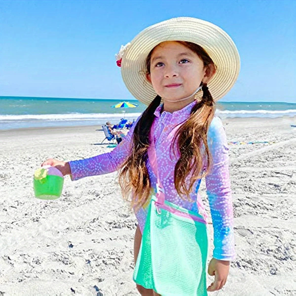 A young girl wearing a straw hat and colorful clothing stands on a beach with the ocean in the background.