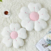 A white plush daisy flower throw pillow resting on a cream-colored fluffy surface with a cup of tea nearby.
