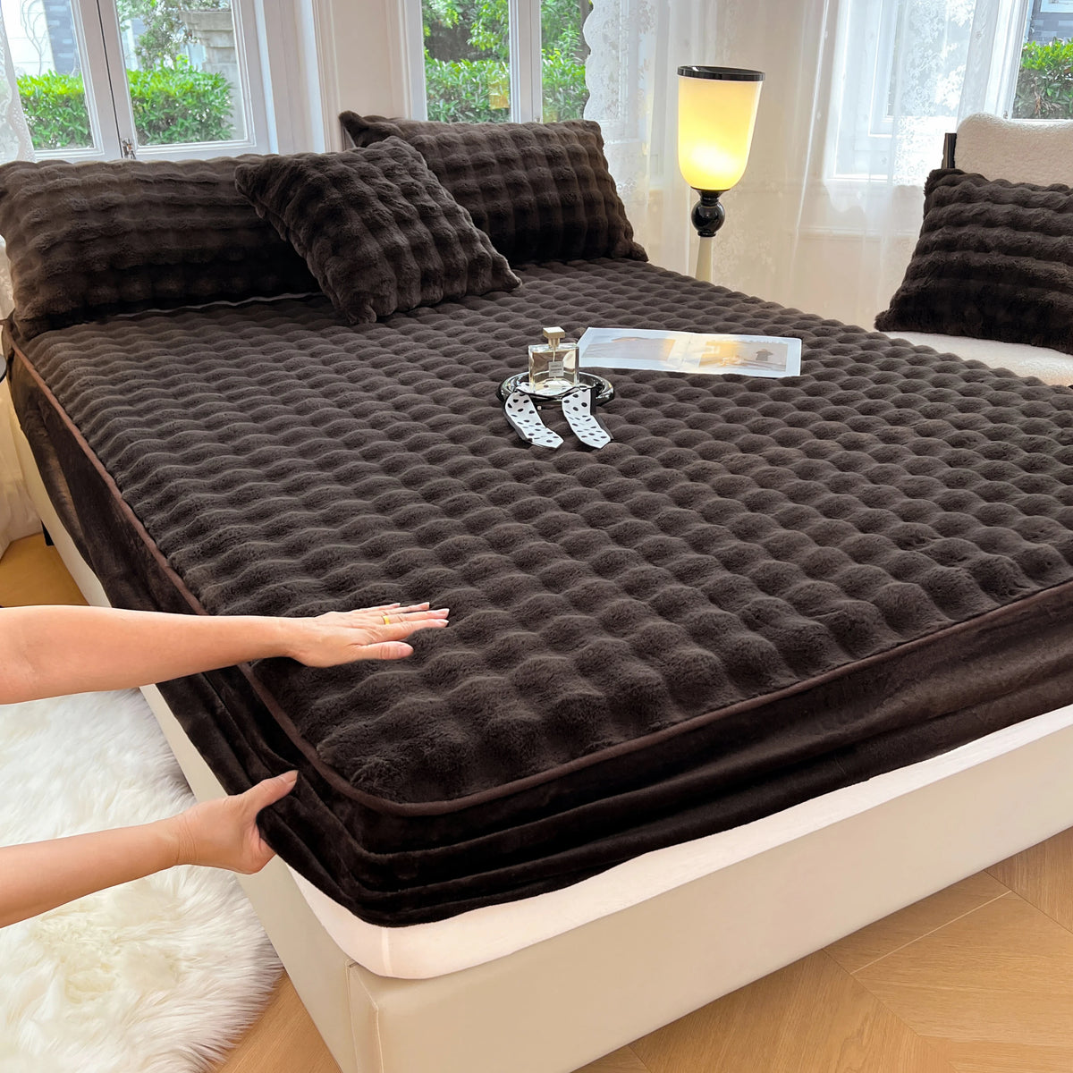 A dark brown fitted sheet set is laid out on a bed, with a hand touching the fabric.