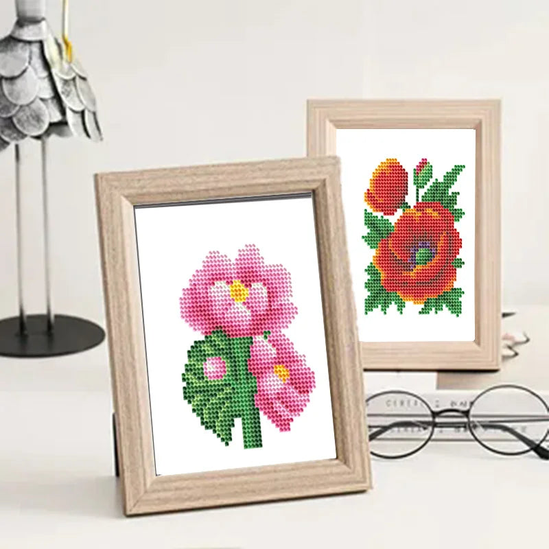 A framed diamond painting featuring a pink flower with green leaves and red center.