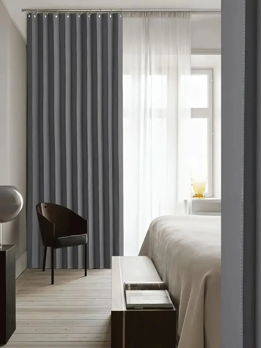 A heavy duty foldable room divider curtain in gray fabric hangs next to a window with sheer white curtains.