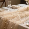 A sheer beige pearl beaded tulle table runner draped elegantly on a white tablecloth.