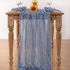 A light blue cheesecloth table runner draped over a wooden table with floral accents for boho wedding decor.