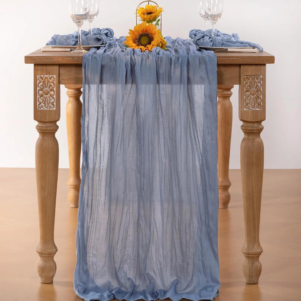 A light blue cheesecloth table runner draped over a wooden table with floral accents for boho wedding decor.