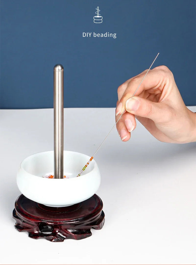 A hand is using a thin stainless steel beading needle to thread beads into a white bowl for DIY beading.