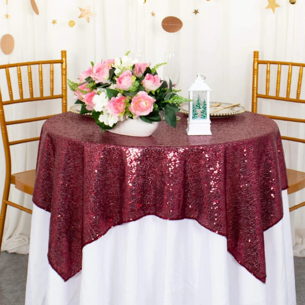 A round table covered with a deep red sequin rectangle tablecloth, decorated with pink and white flowers and a white lantern.