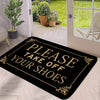 A black doormat with gold lettering that reads Please Take Off Your Shoes