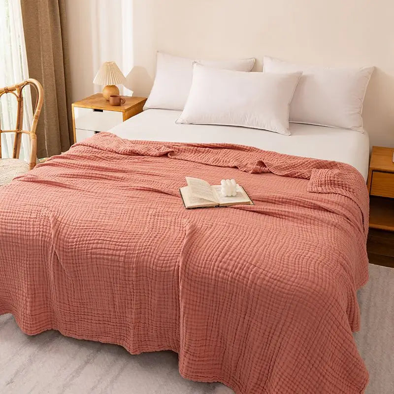 A soft coral pink muslin blanket draped over a white bed with white pillows and natural light.