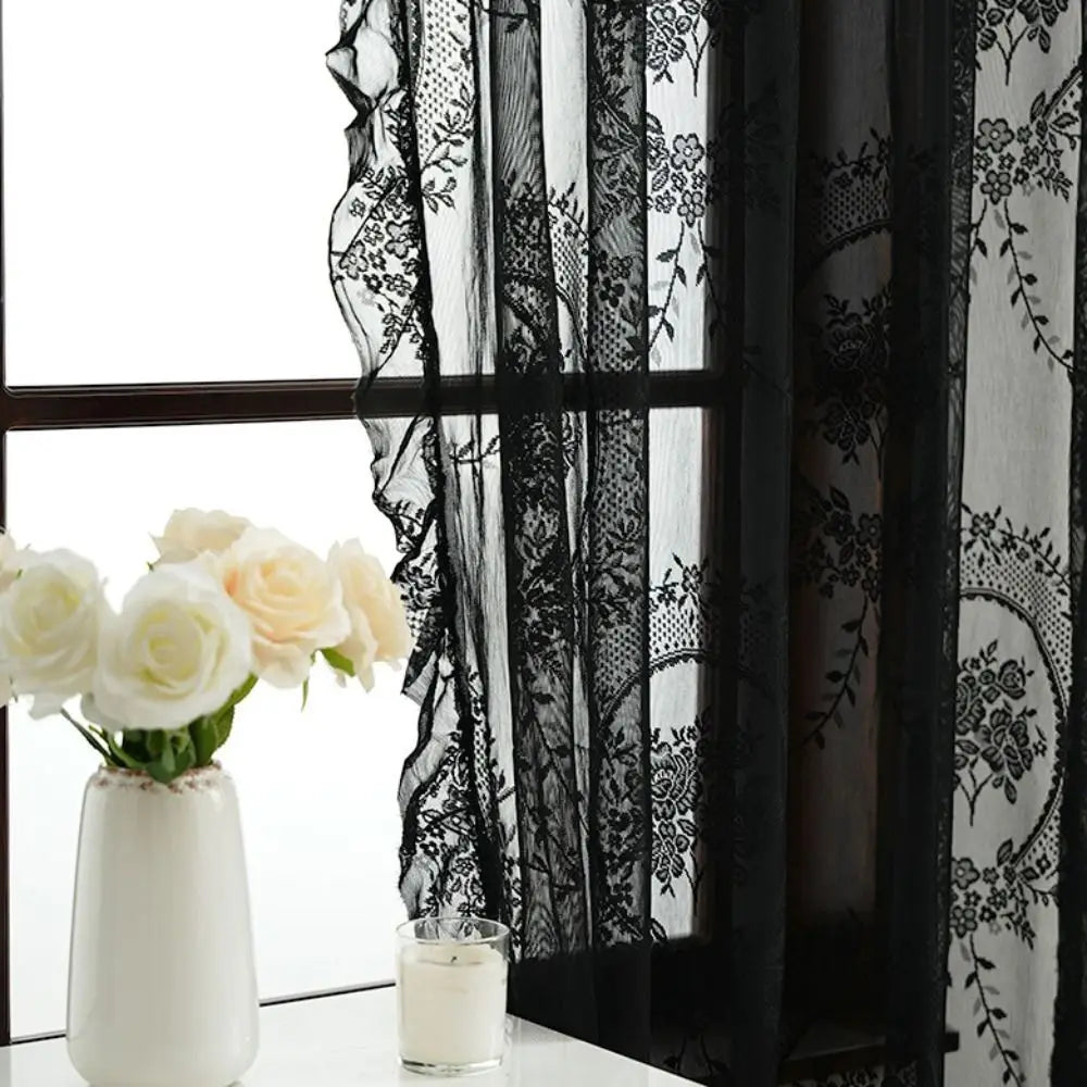 A white ceramic vase holds white roses in front of a black lace and sheer floral curtain set.