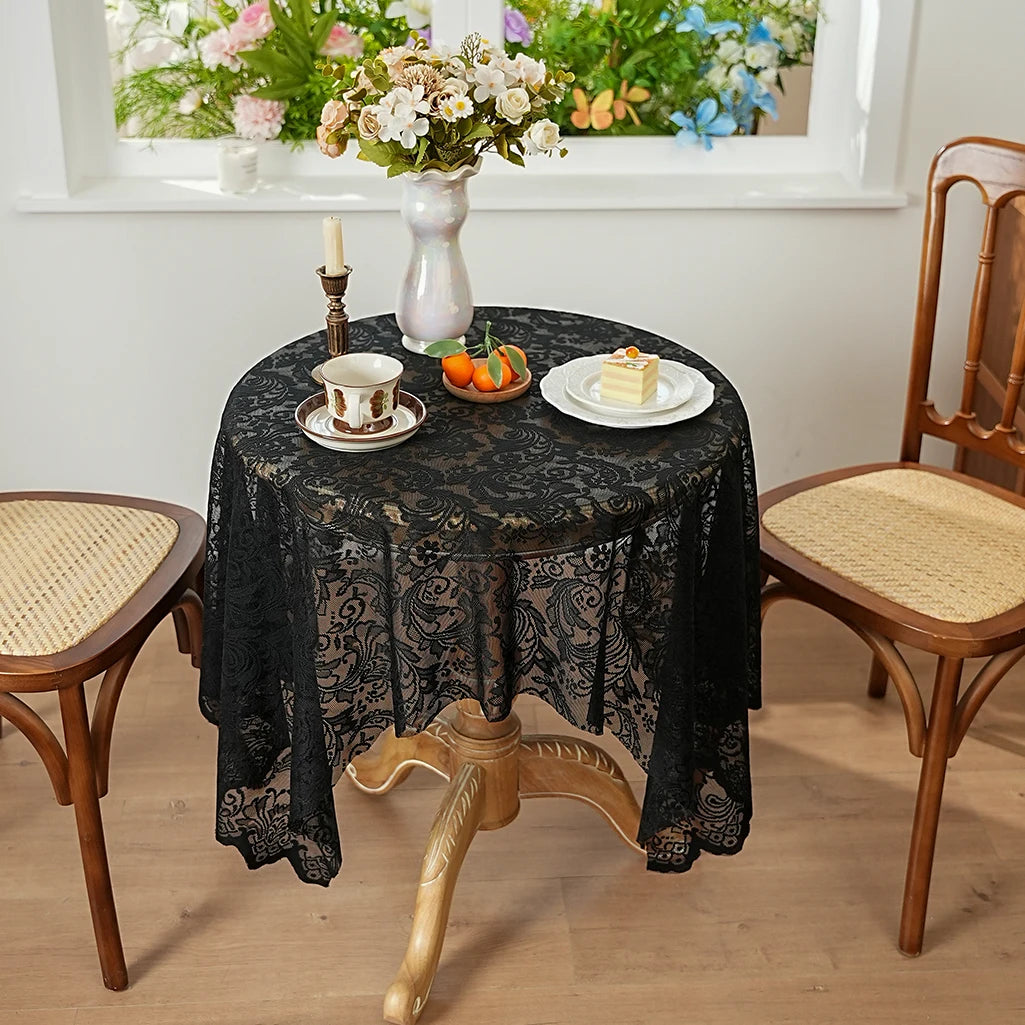 A black lace tablecloth is set on a round table with a cup and saucer, flowers, and a small plate of food.