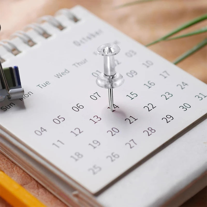 A close-up of a spiral notebook showing a calendar page with push pins and a metal pin.